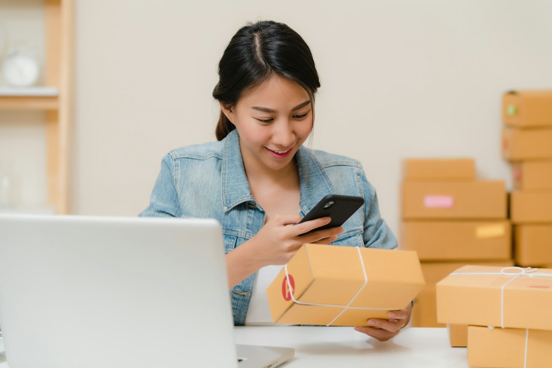 Woman checking package with phone near laptop and boxes - Photo by Rifki Kurniawan on Unsplash