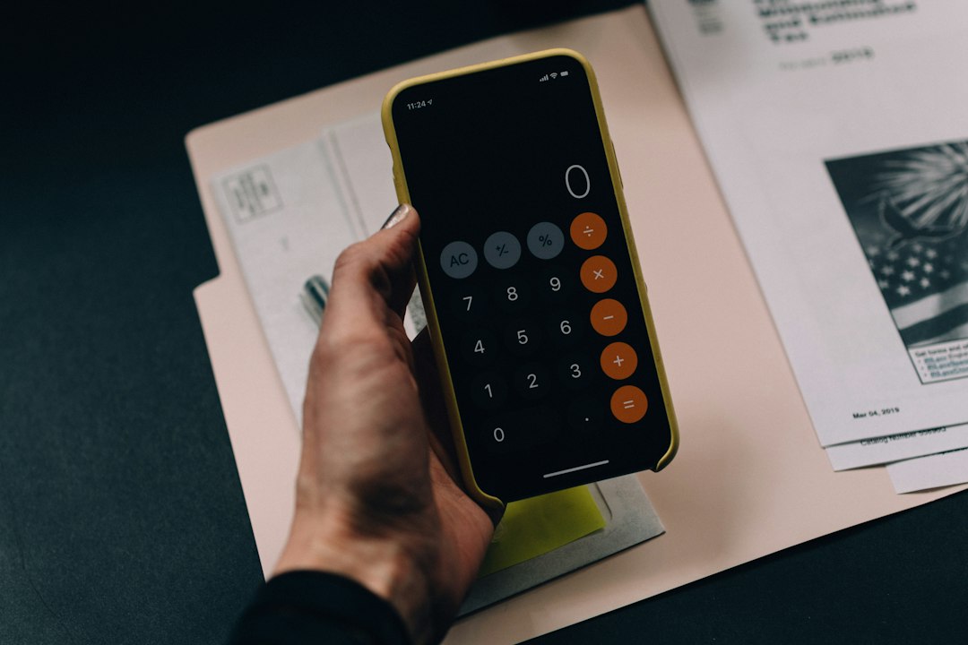 A hand holding a smartphone displaying a calculator app over a paper folder - Photo by Kelly Sikkema on Unsplash